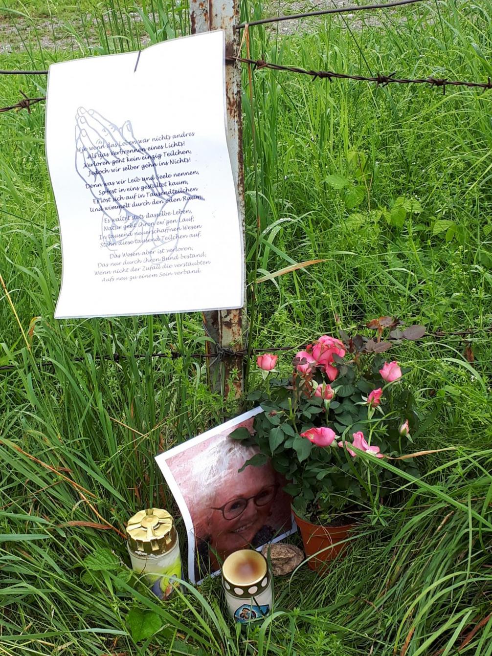 Unfallort von Christine Siess, Stacheldrahtzaun in Wiese mit A4 Zettel (gefaltete Hände und Text) Bild, Kerzen und Blumen